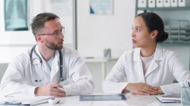 Laboratuvar önlüklü kadın ve erkek doktorlar, tıbbi ofiste masanın başında oturup tartışıyorlar ve sonra kameraya bakıp gülümsüyorlar.
