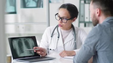 Laboratuvar önlüklü ve gözlüklü genç melez kadın doktor dizüstü bilgisayardaki akciğer röntgenini gösteriyor ve klinikteki tıbbi danışmanlık sırasında erkek hastaya teşhis koymayı açıklıyor.