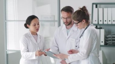 Erkek doktor ve laboratuvar önlüklü iki bayan arkadaşı tıp bürosunda birlikte çalışırken panoda belgeleri tartışıyorlar.