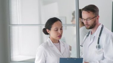 Laboratuvar önlüklü profesyonel erkek ve kadın doktorlar birlikte tıbbi ofiste duruyorlar, panodaki belgeleri tartışıyorlar ve sonra kameraya bakıp gülümsüyorlar.