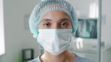 Tek kullanımlık şapka ve koruyucu tıbbi maske takmış genç kadın hemşire portresi hastanede poz verirken kameraya özgüvenle bakıyor. Coronavirüs salgını kavramı