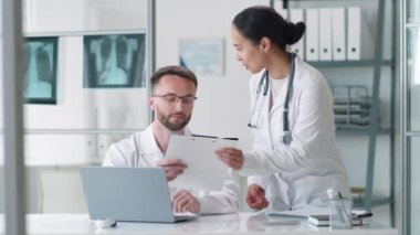 Genç esmer kadın doktor, erkek meslektaşına yürüyor, masaya yaslanıyor, ona pano veriyor ve tıp ofisinde birlikte çalışırken onunla ilgili belgeleri tartışıyor.