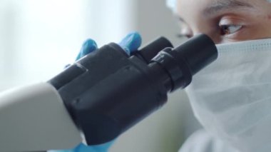 Tıbbi maskeli, tek kullanımlık şapka ve eldiven giyen mikroskop kullanan kadın laboratuvar araştırmacılarının yakın çekimleri.