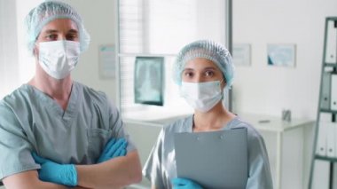 Korunaklı maskeli profesyonel erkek ve kadın doktorlar, tek kullanımlık şapkalar ve eldivenler Coronavirus salgını sırasında hastanede kamera önünde birlikte poz veriyorlar.