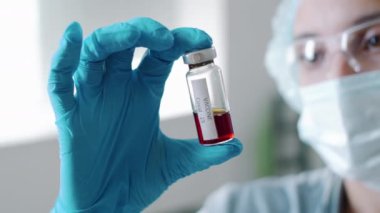 Koruyucu eldivenli, maskeli, şapkalı ve gözlüklü kadın doktorun seçmeli odak noktası. Elinde Covid-19 aşısı olan cam bir şişe var. Hastanede çalışırken sıvıyı inceliyor.