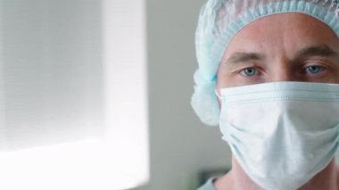 Tek kullanımlık şapkalı ve eldivenli beyaz erkek doktorun portresi tıbbi maske takıyor ve klinikte çalışırken kameraya ciddi şekilde bakıyor. Coronavirüs salgını kavramı