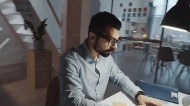 Resmi giyim ve gözlüklü profesyonel Orta Doğulu iş adamının 360 derecelik izleme görüntüsü ofis masasında oturuyor, dizüstü bilgisayarda yazıyor, notlar alıyor ve belgeler okuyor.