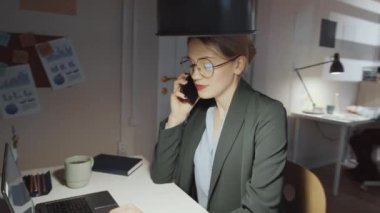 Resmi kıyafet ve gözlüklü genç sarışın iş kadınının ofiste geç saatlere kadar çalışırken masasında oturup konuştuğu 360 derecelik bir çekim.