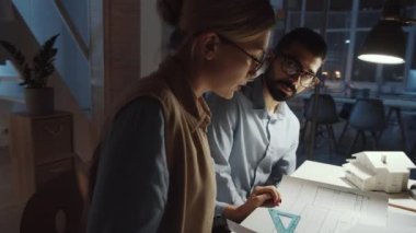 Genç beyaz kadın mimar ve erkek iş arkadaşının ofiste geç saatlere kadar çalışırken masa başında oturup çizimlerini tartışırken 360 derecelik bir izleme görüntüsü.