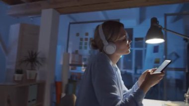 Kablosuz kulaklıklı güzel sarışın kadının gece tasarım stüdyosunda çalışırken zevkle akıllı telefondan müzik dinlediği ve kafasını ritme göre hareket ettirdiği 360 derecelik bir izleme görüntüsü.