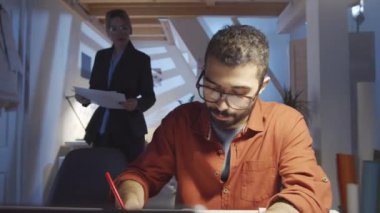 Orta Doğulu erkek tasarımcının çizimlerini 360 derecelik bir açıyla çekerken genç sarışın kadın ona çizimlerle yürüyor. Moda koleksiyonu ve renk paletini tartışan meslektaşlar