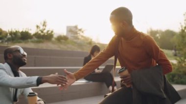 Genç Afrikalı Amerikalı iş adamı parkta kahve ile yürüyor, meslektaşını selamlıyor ve onunla konuşuyor.