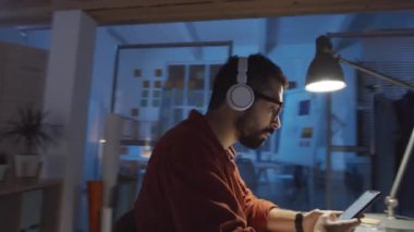 Kablosuz kulaklıklı genç bir adamın gece tasarım stüdyosunda çalışırken akıllı telefondan müzik dinlerken ve şarkı söylerken 360 derecelik izleme görüntüsü.