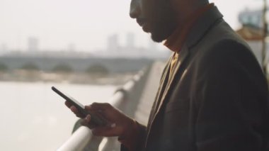 Neşeli Afro-Amerikan iş adamının barajın önünde dikilip akıllı telefondan yazı yazışının orta kesimini kapatın.