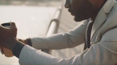 Resmi takım elbiseli dalgın bir Afro-Amerikan işadamı kahveye gitmek için tutunuyor, bariyerlere yaslanıyor ve şehir dışında beklerken mesafeye bakıyor.
