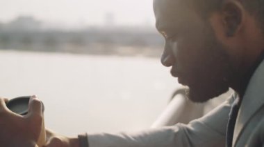 Resmi takım elbiseli Afrikalı Amerikalı işadamı kahve fincanına dalgın dalgın bakıyor ve sonra kentsel setin üzerinde dururken kameraya poz veriyor.