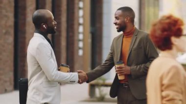İki Afro-Amerikalı iş adamı kahve fincanlarına sarılıp el sıkışarak selamlaşıyorlar ve sokakta konuşuyorlar.