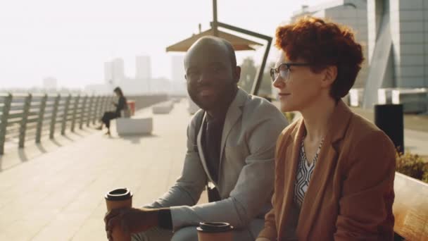 アフリカ系アメリカ人の実業家と白人の実業家がコーヒーカップを持ち 街の堤防のベンチに座りながら話をする — ストック動画