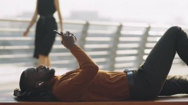 Kablosuz kulaklıklı genç Afrikalı Amerikalı bir adam ve şehirde bankta uzanmış akıllı telefon kullanıyor.