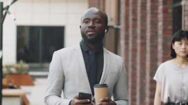 Resmi takım elbiseli Afro-Amerikan iş adamı şehirde sokakta yürüyor, kahve içmeye gidiyor ve cep telefonundan kablosuz kulaklıklar aracılığıyla konuşuyor.
