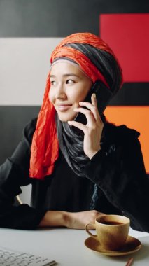 Orta boy, tesettürlü İslami Ortadoğulu genç bir kadının dikey görüntüsü. Ofiste oturuyor, cep telefonuyla gülümsüyor.