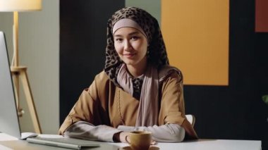 Orta ölçekli, genç Müslüman Orta Doğulu kadın tesettüre oturmuş, gündüz kameraya bakıp gülümsüyor.