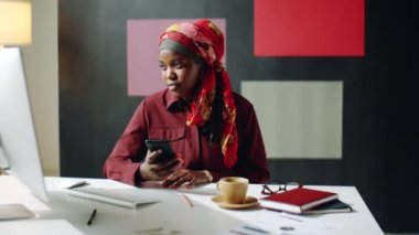 Renkli başörtüsü takan, ofisinde oturan, akıllı telefon kullanan siyahi bir kadının orta boy yakın çekimi.