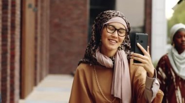 Genç mutlu Orta Doğulu kadın tesettür ve gözlük takıyor. Gündüz vakti akıllı telefondan arama yapıyor.