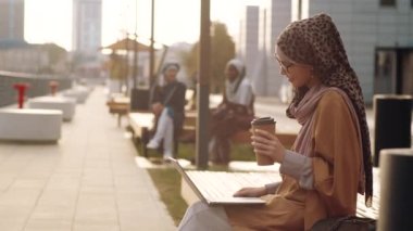 Orta ölçekli, tesettürlü ve gözlüklü genç bir Orta Doğulu kadının yaya kaldırımındaki bankta oturması dizüstü bilgisayarla video araması.