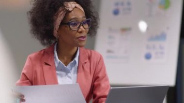 Afrika kökenli Amerikalı iş kadını elinde belge tutuyor ve genç erkek meslektaşıyla ofis toplantısında projeyi tartışıyor.