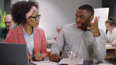 Afrika kökenli Amerikalı erkek ve kadın meslektaşlar ofis masasında birlikte oturup iş projelerini iş günlerinde dizüstü bilgisayarlarında konuşurken gülümsüyorlar.