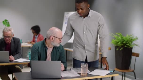若いアフリカ系アメリカ人の男性が机の上にもたれかかり オープンスペースのオフィスで働いている間 ラップトップやシニア白人同僚への書類に関するプロジェクトについてアドバイスをする — ストック動画