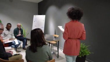 Afro-Amerikan kadın iş koçu seminer odasına giriyor, pano ile duruyor ve karışık yaştaki farklı çalışanlardan oluşan bir ekiple konuşuyor ve onu alkışlıyorlar.