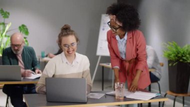 Neşeli Afrikalı Amerikalı bayan yönetici, genç beyaz bir kadına doğru yürüyor, masaya yaslanıyor ve onunla dizüstü bilgisayardaki projeyi tartışıyor ve aynı zamanda açık uzay ofisinde çalışanlarla birlikte çalışıyor.