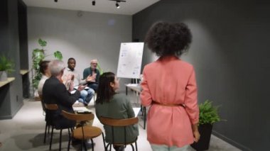 Afrika kökenli Amerikalı kadın işletme koçu seminer odasına giriyor, karışık etnik gruplardan oluşan bir grup çalışanı selamlıyor ve gülümsüyor.