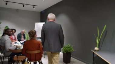 Kıdemli erkek işletme koçu seminer odasına girerken çok ırklı bir grup onu alkışlıyor, sonra masanın yanında duruyor ve seyircilere bir konuşma yapıyor.
