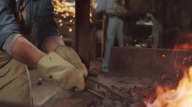 Önlüklü ve koruyucu eldivenli sakallı demircinin atölyedeki ocağı yakmak için metal çubuk kullanırken çekimini yap.
