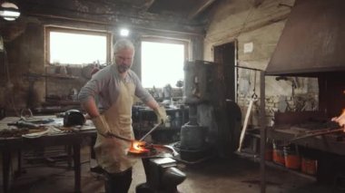 Önlük ve koruyucu eldivenler üzerinde deneyimli demirci. Atölyede iş arkadaşlarıyla çalışırken ısıtılmış demir çubuk şekillendiriyor.