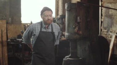Genç sakallı demirci atölyede yürüyor, el sıkışıyor ve kıdemli meslektaşıyla konuşuyor.