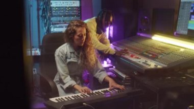 Genç beyaz kadın, kayıt stüdyosunda çalışırken karıştırma konsolu kullanan sentezleyici ve Afrikalı Amerikalı erkeği oynuyor.