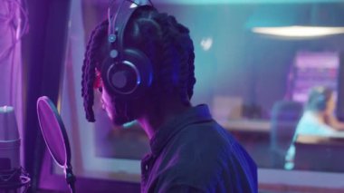 Kulaklıklı havalı Afro-Amerikalı adamın ses prodüksiyon stüdyosunda rap şarkısı kaydedişinin yan görüntüsü
