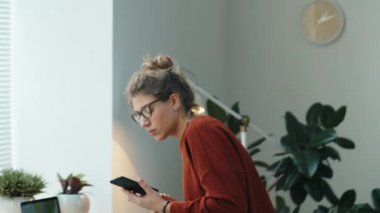 Akıllı telefon kullanan genç bir iş kadını, sonra ofis masasında oturup, dizüstü bilgisayardaki interneti karıştırıyor ve not alıyor.