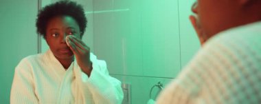 Genç Afrikalı Amerikalı kadın pamuklu ped ile yüz toneri uyguluyor ve banyo aynasına bakıyor.