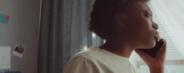 Genç Afrikalı Amerikalı bir kadının evinin penceresinde durup cep telefonuyla sohbet ederken ve gülümserken yakın plan çekimleri.