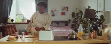Genç Afrikalı Amerikalı kadın ottan kahve dolduruyor, içiyor ve sonra mutfakta kahvaltı ederken tost makinesinden bir dilim ekmek alıyor.