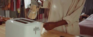 Genç Afrikalı Amerikalı kadın kahve içiyor ve kahvaltıda tost makinesinden bir dilim ekmek alıyor.
