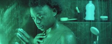 Genç Afrikalı Amerikalı kadın duş kabininde durmuş, yukarıdan su akıyor, ıslak cam duvarda el ele tutuşuyor ve dalgın dalgın bakıyor.