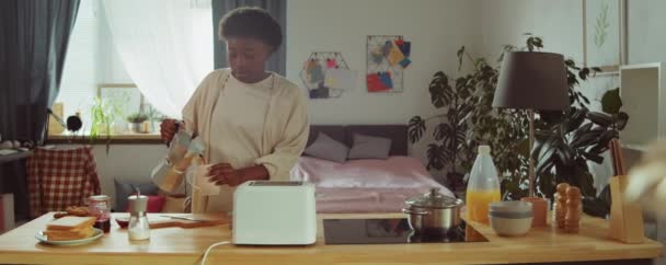 年轻的非洲裔美国女人把咖啡倒入锅中 然后在厨房吃早饭时从烤面包机上取下一片面包 — 图库视频影像