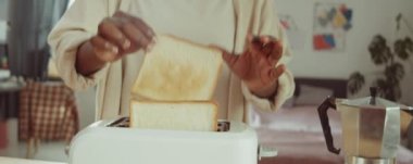 Genç Afrikalı Amerikalı kadın tost makinesinden bir dilim ekmek alıyor ve evde kahvaltı hazırlarken üzerine reçel koyuyor.