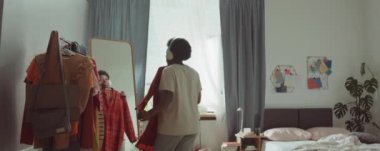 Kablosuz kulaklıkla dans eden ve yatak odasında aynanın önünde kıyafet seçen genç Afrikalı Amerikalı kadını yakınlaştırın.